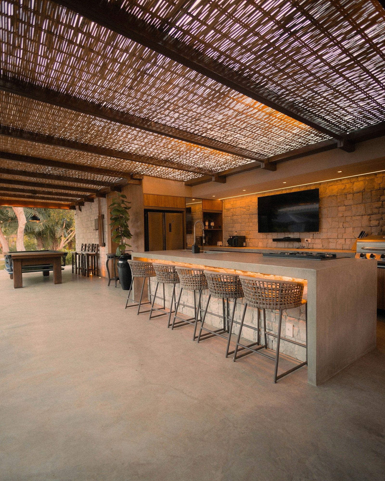 A long concrete-topped outdoor kitchen island with woven bar stools, warm under-counter lighting and a stone feature wall beneath a timber pergola.