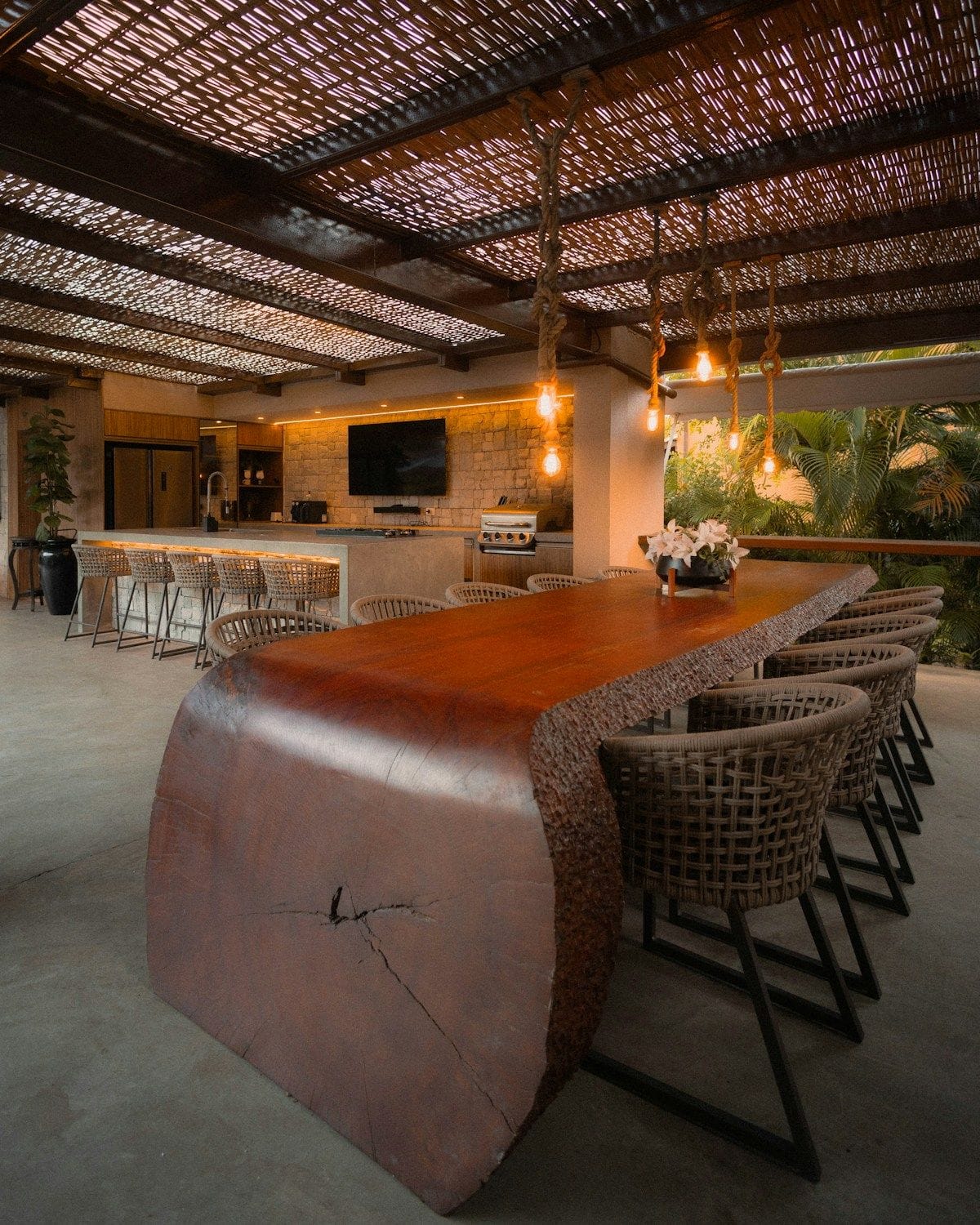 A live-edge timber bar top with stools inside a covered outdoor kitchen, lit by pendant lights.